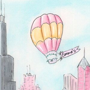 May include: A pink and yellow hot air balloon with a banner that says "Emma's" is flying over a city skyline. The buildings are gray, pink, and white.