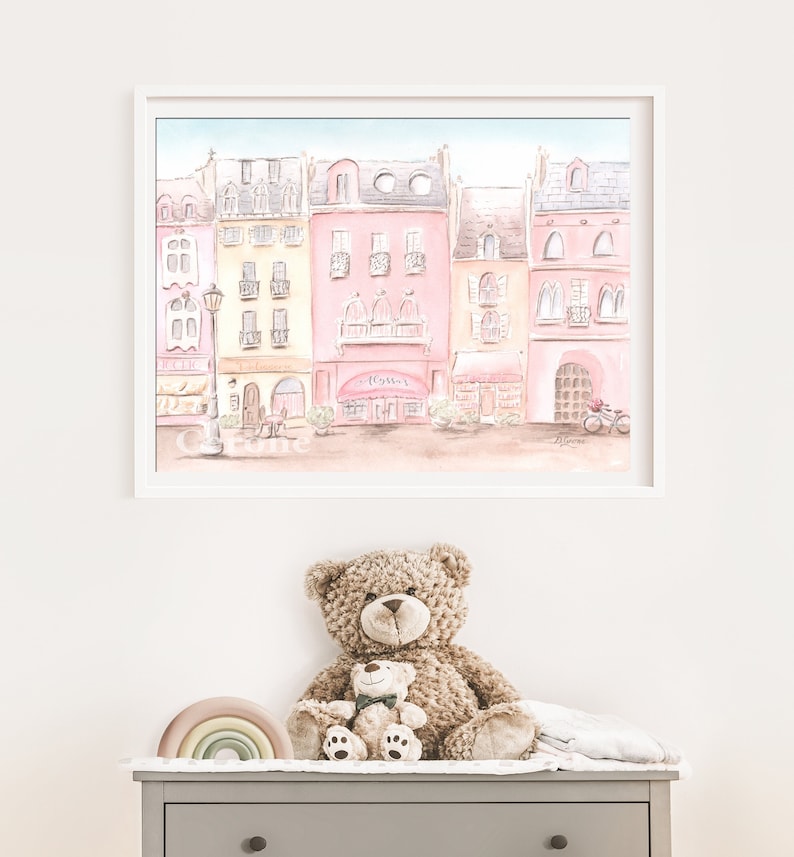 Op de afbeelding: Een aquarel schilderij van een rij pastelroze en gele gebouwen met een caf&eacute; genaamd "Tom's" in het midden. Het schilderij is wit ingelijst en hangt aan een witte muur. Een bruine teddybeer zit op een grijze commode met een regenboogkleurig houten speelgoed ervoor.