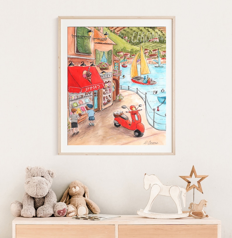 Op de afbeelding: Een aquarel schilderij van een kleurrijk Italiaans kustplaatsje met een rode scooter, een bakkerij genaamd "Emma's" en twee kinderen die langs lopen. De sc&egrave;ne bevat een zeilboot op het water en een groene heuvel op de achtergrond.