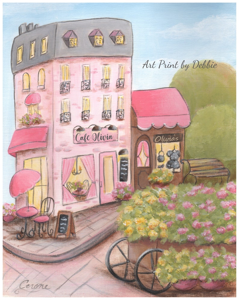 Op de afbeelding: Een kunstafdruk van een roze Parijs caf&eacute;, "Cafe Olivia", met een grijs dak en roze luifels. Een bloemenkar en zitplaatsen buiten staan ervoor. De afdruk is gesigneerd "Cerone" en zegt "Art Print by Debbie".