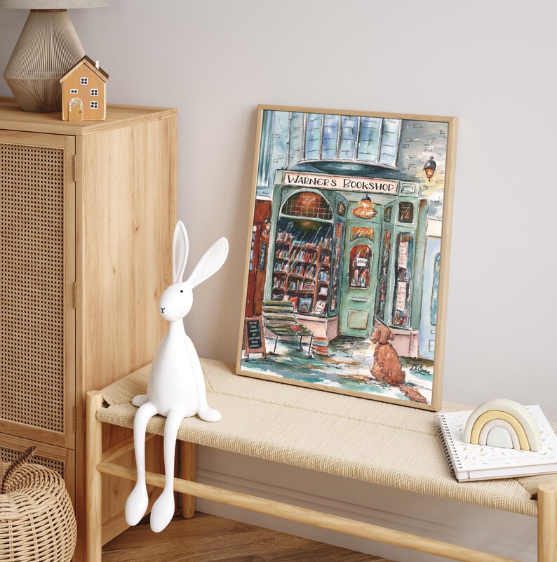 May include: A watercolor painting of a bookstore with a green awning and the words "Warner's Bookshop" above the entrance. A dog sits outside the shop, looking at the door. The painting is framed in a light wood frame.