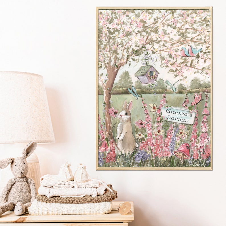 K&ouml;nnte beinhalten: Ein gerahmter Aquarelldruck mit einer Gartenszene mit einem Hasen, Blumen und einem Vogelhaus. Der Druck enth&auml;lt den Text "Gianna's Garden" auf einem Schild. Das Kunstwerk befindet sich in einem Holzrahmen und wird an einer wei&szlig;en Wand ausgestellt.