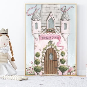 K&ouml;nnte beinhalten: Ein gerahmter Aquarelldruck eines rosa Schlosses mit dem Text "Princess Olivia" auf einem Banner. Das Schloss hat eine Holzt&uuml;r, gew&ouml;lbte Fenster und ist von Gr&uuml;n und Blumen umgeben. Eine Puppe mit einer Krone befindet sich im Vordergrund.