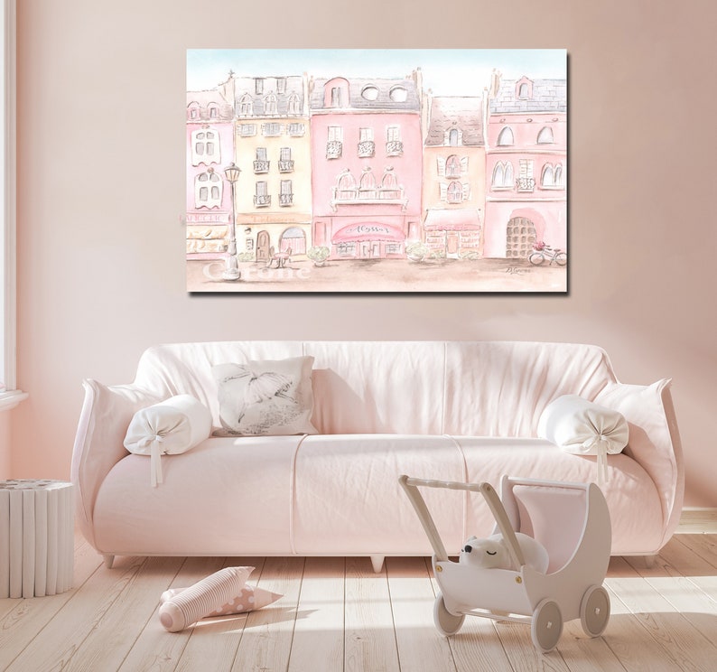 Op de afbeelding: Een aquarel schilderij van een rij pastelkleurige gebouwen met een lantaarnpaal en een fiets ervoor. De gebouwen hebben ramen en deuren, en sommige hebben luifels. Het schilderij is in een lichtroze lijst en hangt aan een roze muur.