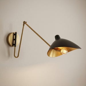 May include: A wall-mounted, adjustable lamp with a black shade and a gold interior. The lamp features a brass arm and a circular wall mount. The light bulb is visible inside the shade, illuminating the gold interior.