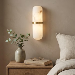 May include: A modern wall sconce with a white marble-like finish and a brass band. The elongated, capsule-shaped light fixture is mounted on a neutral-colored wall. A nightstand with a vase, book, and glasses is in the foreground.