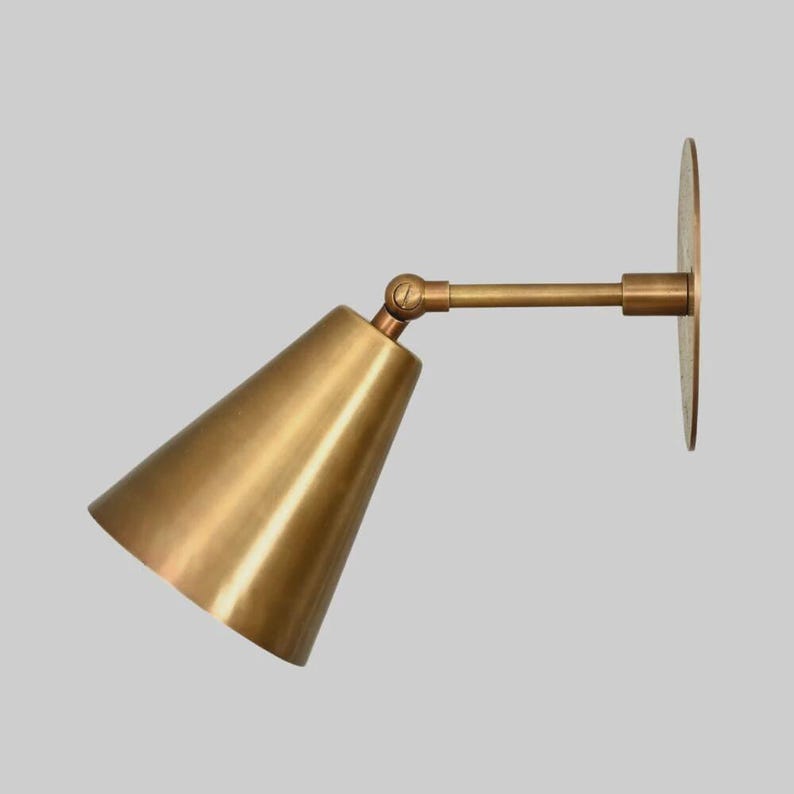 May include: A brass wall-mounted sconce with a conical shade and adjustable arm. The sconce features a circular mounting plate and a warm, metallic finish. The design is minimalist and suitable for various interior styles.