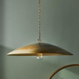 May include: A brass-coloured pendant light with a wide, shallow shade. The shade has a slightly domed shape and is suspended by a chain. A single light bulb hangs from the centre of the shade. The background is a muted green.