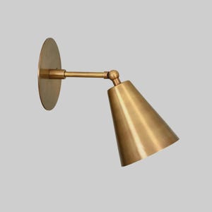 May include: A brass wall-mounted sconce with a circular backplate, an adjustable arm, and a conical shade. The fixture is a warm gold color and is designed to direct light downwards. The design is minimalist and modern.