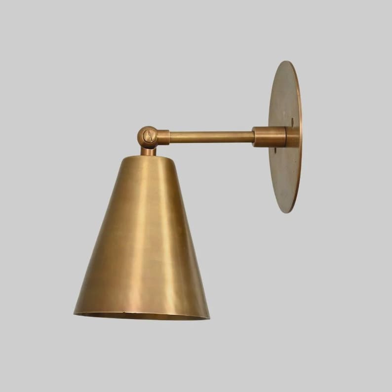 May include: A brass-toned wall sconce with a conical shade and a circular backplate. The sconce features an adjustable arm and a warm, metallic finish, suitable for interior lighting. The design is minimalist and modern.