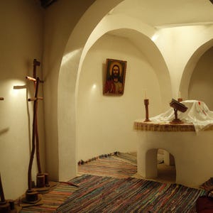May include: A white arched interior space featuring two tall wooden crosses on the left, a framed religious portrait on the wall, and a central white altar covered with a white cloth, holding a small book and two thin candles on wooden stands.