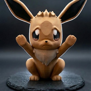 Op de afbeelding: Een 3D-geprint Eevee-figuur in een zittende pose, met de armen omhoog. Het figuur is voornamelijk lichtbruin met zwarte accenten op de oren. Het heeft grote, expressieve ogen en een witte, vachtachtige kraag. Geplaatst op een donker, getextureerd oppervlak.