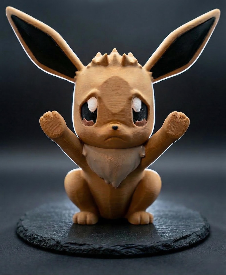 Op de afbeelding: Een 3D-geprint Eevee-figuur in een zittende pose, met de armen omhoog. Het figuur is voornamelijk lichtbruin met zwarte accenten op de oren. Het heeft grote, expressieve ogen en een witte, vachtachtige kraag. Geplaatst op een donker, getextureerd oppervlak.