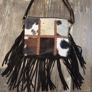 May include: A patchwork crossbody bag with a cowhide front, featuring squares of black, white, and brown fur. The bag has a black fringe along the bottom and a brown leather strap. The bag is designed with a zipper closure and gold-colored hardware.