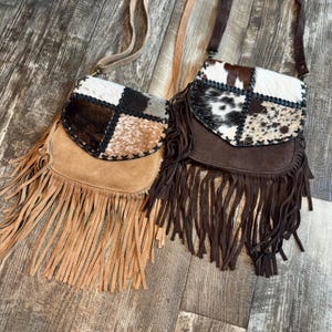 May include: Two crossbody bags with long fringe detailing. The left bag is tan suede with a cowhide flap featuring brown, black, and white patches. The right bag is dark brown suede with a cowhide flap featuring black and white spotted patterns.