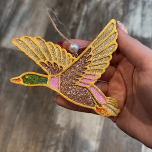May include: A decorative ornament in the shape of a flying duck. The duck has gold glitter wings, a brown glitter body, and pink and green accents. It is suspended by a twine string and a pearl bead.