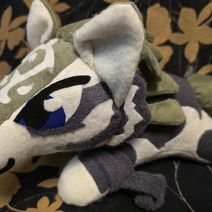 May include: A plush toy in the shape of a stylized animal, featuring a white, gray, and green color scheme. The toy has a detailed design with patterned accents, including a blue eye and a black outline. The plush is soft and cuddly.