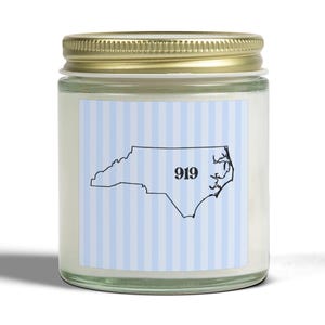 May include: A glass jar candle with a gold lid. The label features a blue and white striped background with a black outline of North Carolina and the number "919". The candle sits on a white surface.