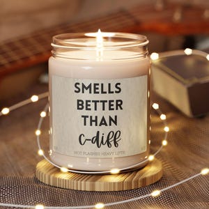 May include: A lit candle in a glass jar with a label that reads "Smells Better Than c-diff". The candle sits on a small wooden coaster, with string lights and a guitar in the background. The text "Hot Flashes Heavy Lifts" is below the main text.