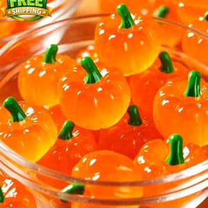 May include: A clear glass bowl filled with multiple small, translucent orange pumpkin-shaped sweets. Each sweet features a smooth, glossy surface with vertical ridges and a distinct, solid green stem protruding from the top centre of the pumpkin.
