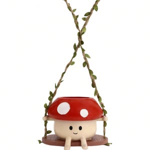 Peut inclure: Un pot de fleurs en forme de champignon avec un chapeau rouge et des taches blanches, suspendu par de la ficelle ornée de feuilles vertes. Le pot a un visage souriant et est conçu pour les petites plantes ou fleurs. La base est marron.