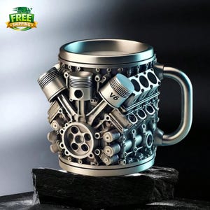 May include: A silver-coloured mug shaped like a V6 engine, with pistons and engine components visible. The mug has a handle and sits on a dark, textured surface. The text "V6" is visible on one of the pistons.