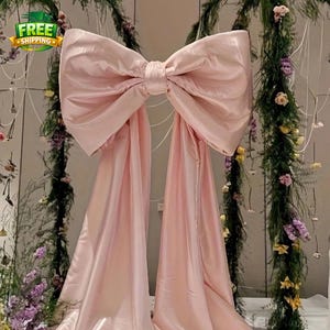 May include: A large, light pink satin bow with long, flowing tails. The bow is the focal point, set against a backdrop of greenery and flowers, suggesting a wedding or event decoration. The bow is centered and appears to be made of a soft, draping fabric.