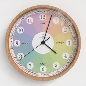May include: A round wall clock with a light wood frame and a multicolored face. The dial features numbers 1 through 12 in white circles, an outer ring with minutes 1 through 60, and text labels o'clock, quarter past, half past, and quarter to.