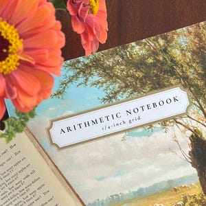 May include: An arithmetic notebook with a 1/4-inch grid, featuring a cover illustration of a pastoral landscape with trees and grazing cows. The open notebook displays printed math word problems on the left page, with vibrant pink and orange zinnia flowers positioned in the upper left corner.