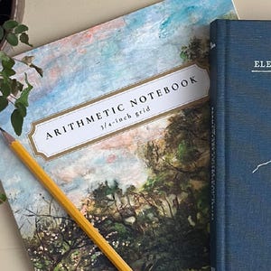 May include: An arithmetic notebook with a 3/4-inch grid, featuring a landscape painting cover with trees and a blue sky. A yellow pencil with a pink eraser rests diagonally across the notebook, next to a dark blue hardcover book titled Elementary.