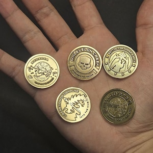 Puede incluir: Cinco monedas doradas con varios diseños y texto. Una moneda dice "MERIDIA" con un gráfico. Otra dice "MALEVELON CREEK" con una calavera. Otras monedas tienen texto como "BATTLE FOR SUPER EARTH" y "LIBERATION OF OSHAUNE".