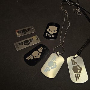Könnte beinhalten: Eine Sammlung von Erkennungsmarken aus Metall in Schwarz und Silber mit eingravierten Totenkopf- und Flügelmotiven. Einige Marken tragen den Schriftzug SEAF. Zwei Marken sind an Kugelkettchen befestigt, während andere lose mit verschiedenen Designs liegen.