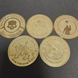 May include: Five circular gold-colored metal coins featuring embossed designs and text. The coins display various emblems and phrases including Malevelon Creek, Meridia, Battle for Super Earth, Liberation of Oshaune Into the Hive, and Helldivers Battle for Cyberstan.