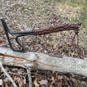 May include: A black metal hatchet with a wrapped handle in a pattern of red, orange, and black. The hatchet is resting on a tree branch, showcasing its design and craftsmanship. A loop of cord is attached to the handle.