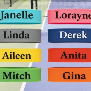 May include: A selection of vibrant wristbands, each printed with a name in a bold black typeface. The bands are in a range of colours: teal, pink, grey, blue, yellow, red, green, and orange. Names include Janelle, Lorayne, Linda, Derek, Aileen, Anita, Mitch, and Gina.