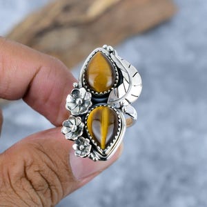 May include: A silver ring featuring two teardrop-shaped tiger's eye stones. The ring has a floral and leaf design with small, detailed flowers and a textured leaf. The stones are a warm, golden-brown color, set against the silver metal.