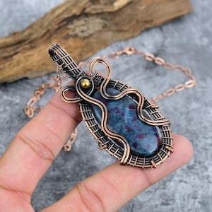 Puede incluir: Collar con colgante de cobre hecho a mano con una piedra azul y roja. La piedra está encerrada en un diseño de alambre de cobre tejido, con una cadena de cobre. El colgante tiene forma ovalada.