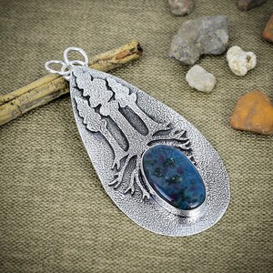 May include: A teardrop-shaped silver pendant featuring a textured surface with a raised tree design and a single oval-shaped blue and green gemstone set in the centre. The pendant includes two silver loops at the top for attachment to a chain.