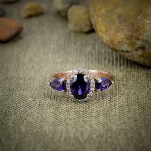 Könnte beinhalten: Ein Ring aus Roségold mit einem zentralen, oval geschliffenen, tiefvioletten Edelstein, der von einem Kranz aus kleinen weißen Steinen umgeben ist. Zwei tropfenförmige violette Edelsteine flankieren den zentralen ovalen Stein.