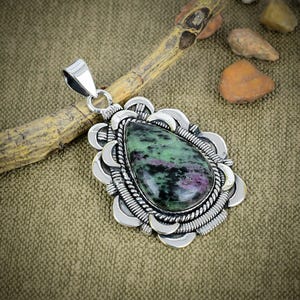 Puede incluir: Un colgante en forma de lágrima con una piedra de zoisita pulida que presenta inclusiones verdes, negras y magenta. La piedra está engastada en un marco de metal plateado decorativo con detalles de cuerda y bordes festoneados.