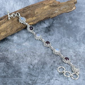 May include: A silver chain bracelet featuring alternating round cabochon moonstones and dark red garnet gemstones. Each stone is set in a decorative scalloped silver bezel. The bracelet includes a toggle clasp closure with three circular silver links.
