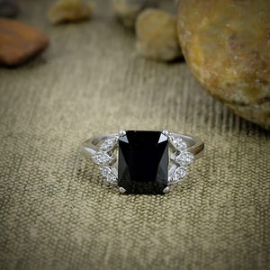 May include: A silver-toned ring featuring a large, rectangular, faceted black center stone. The band is accented on both sides by a cluster of three small, clear, round-cut stones arranged in a leaf-like pattern, set in a polished metal frame.