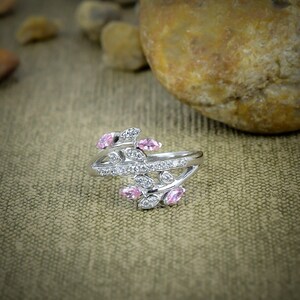 May include: A silver ring with a leaf design, featuring pink and clear gemstones. The ring has a split shank and is set with small, round, and oval-shaped stones. The gemstones are arranged to resemble leaves and branches.