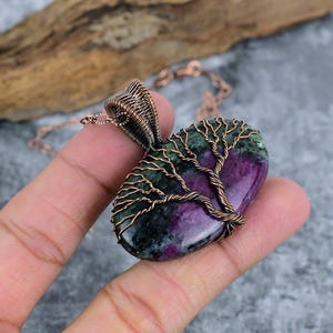 Puede incluir: Un collar con colgante envuelto en alambre de color cobre con una piedra pulida de forma ovalada. La piedra tiene una combinación de colores púrpura y verde intenso, con un diseño de árbol de alambre superpuesto a la piedra.