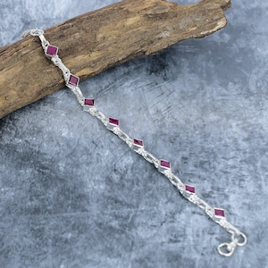 May include: A silver chain bracelet featuring seven evenly spaced, square-cut, deep pinkish-red gemstones set in polished silver frames. The bracelet has a delicate link design and a small circular clasp at one end, showcasing a refined aesthetic.