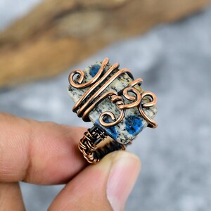May include: A handcrafted ring with a rectangular stone displaying blue and cream patterns, encased in copper wire. The wirework incorporates decorative swirls and wraps around the stone and the ring band. The ring is set against a blurred backdrop.