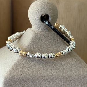 May include: A stretch bracelet composed of alternating polished silver-tone and gold-tone spherical beads of varying sizes. The beads are arranged in a repeating pattern along the circular band, creating a metallic finish with reflective surfaces.