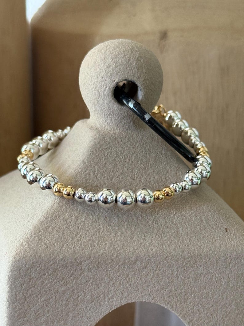 May include: A stretch bracelet composed of alternating polished silver-tone and gold-tone spherical beads of varying sizes. The beads are arranged in a repeating pattern along the circular band, creating a metallic finish with reflective surfaces.