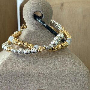 May include: A multi-strand beaded bracelet featuring a mix of polished gold-tone, silver-tone, and smooth white spherical beads. The beads are arranged in a repeating pattern along the elastic cord, creating a layered look with metallic finishes.