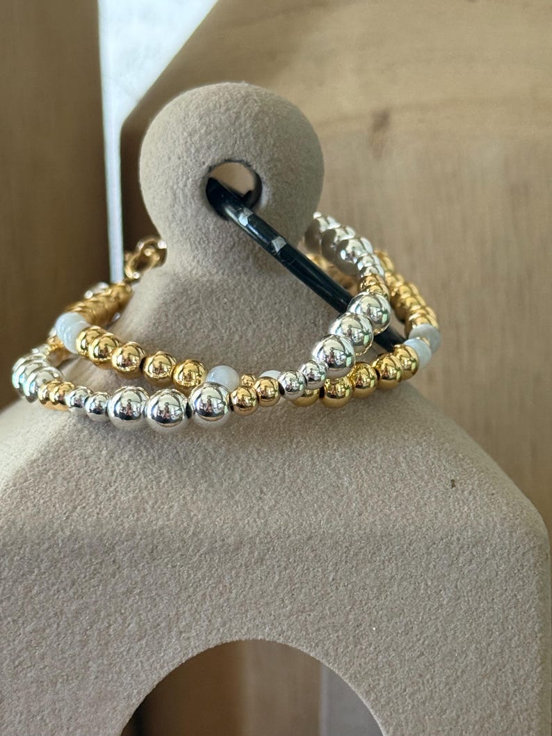 May include: A multi-strand beaded bracelet featuring a mix of polished gold-tone, silver-tone, and smooth white spherical beads. The beads are arranged in a repeating pattern along the elastic cord, creating a layered look with metallic finishes.